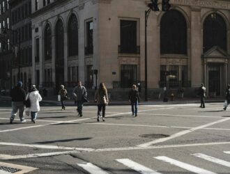People walking across a crosswalk in downtown Hartford near a historic stone building on a sunny day.