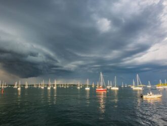 Sailboats anchored in a harbor under dark storm clouds, reflecting environmental uncertainty and climate impact on communities.