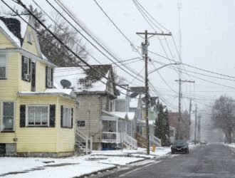 Snow-covered residential street with houses, power lines, and cars, showing everyday community infrastructure during winter conditions.