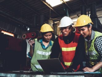 Three workers in hard hats and safety vests reviewing work on a laptop in a factory.